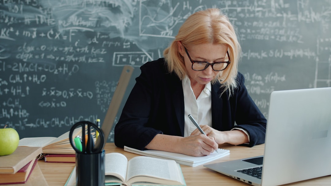 Mature woman in suit working in class writing sitting at desk with books and laptop, blackboard with formulas is visible in background. People and work concept.